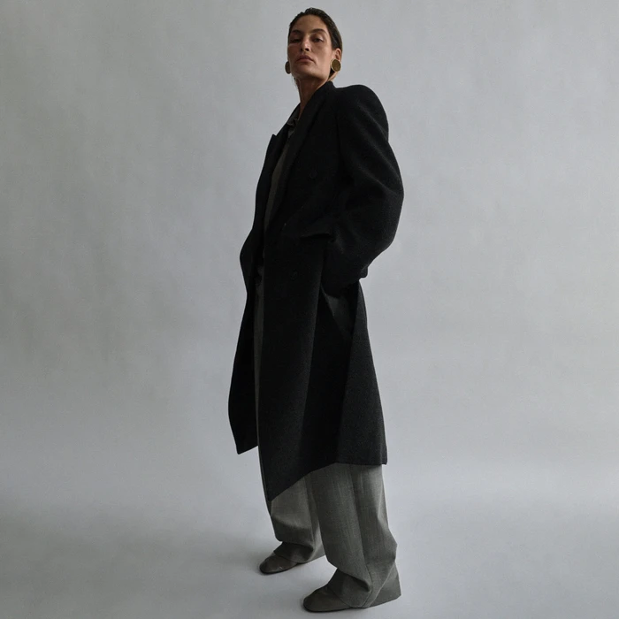 Model in a black coat and grey trousers standing against a pale grey background for Phoebe Philo