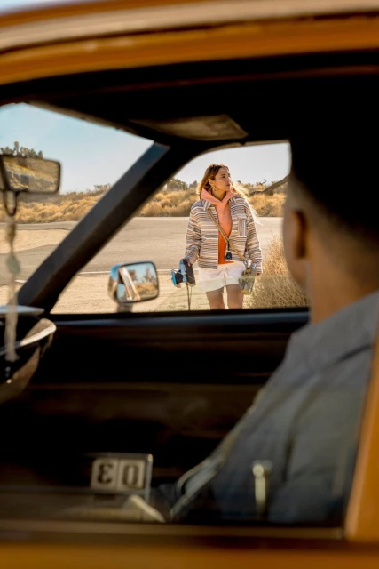 A woman standing near an orange car on a desert road is seen from the inside of the car, looking at the woman from the driver's seat.