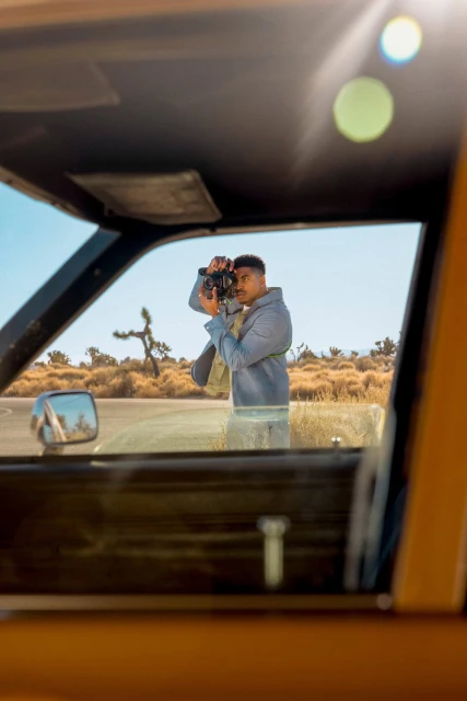 Photographer with camera taking pictures in desert landscape, seen framed through the window of a vintage car on a sunny day.