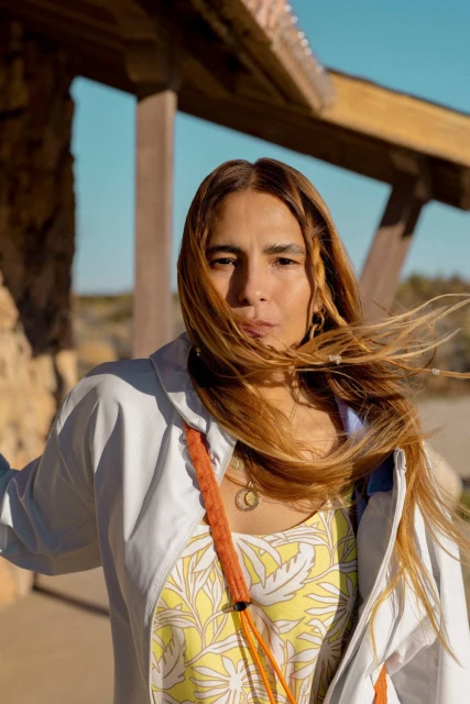 Portrait of a person with long brown hair standing outside wearing a light jacket, and a yellow patterned top on a sunny day.