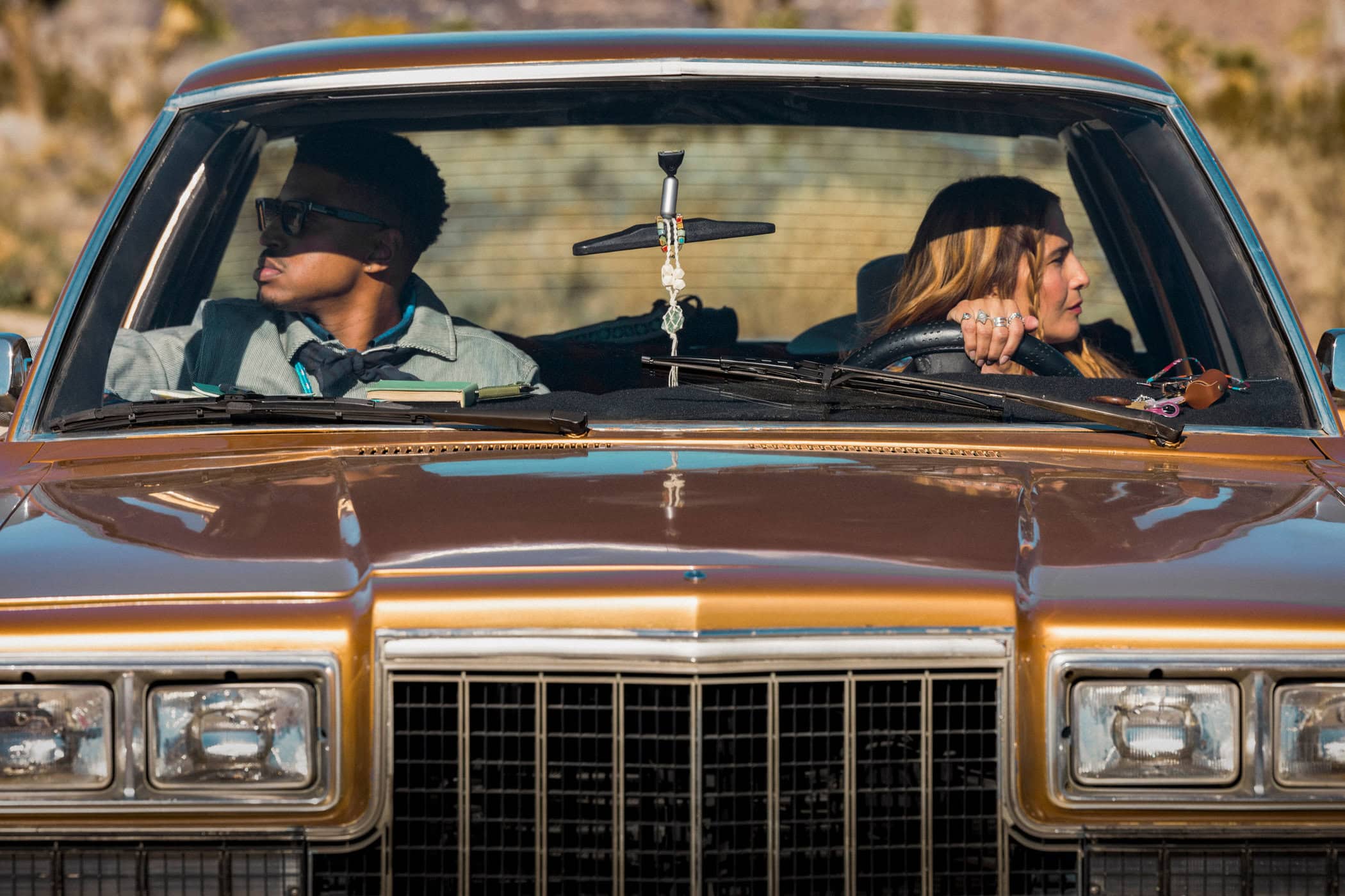 Two people, an adult male and an adult female, sit inside a classic brown car on what looks like an outdoor road trip in daytime.