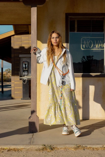 Woman in a dress stands near a roadside building with vintage details in an outdoor desert landscape under the sun.