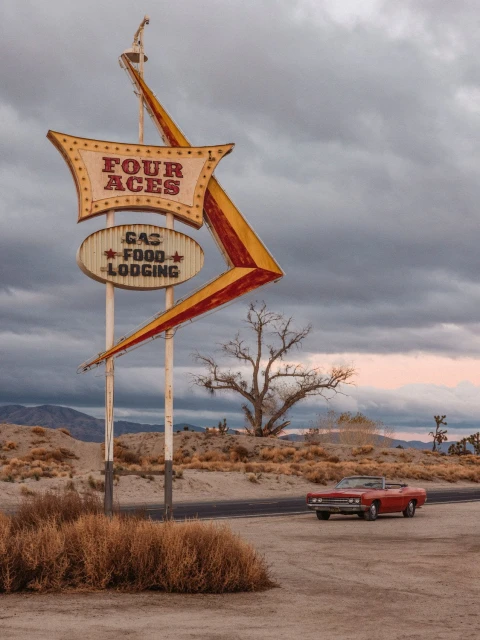 Vintage Four Aces motel road sign along desert highway, set against an overcast sky with a classic red convertible car nearby.