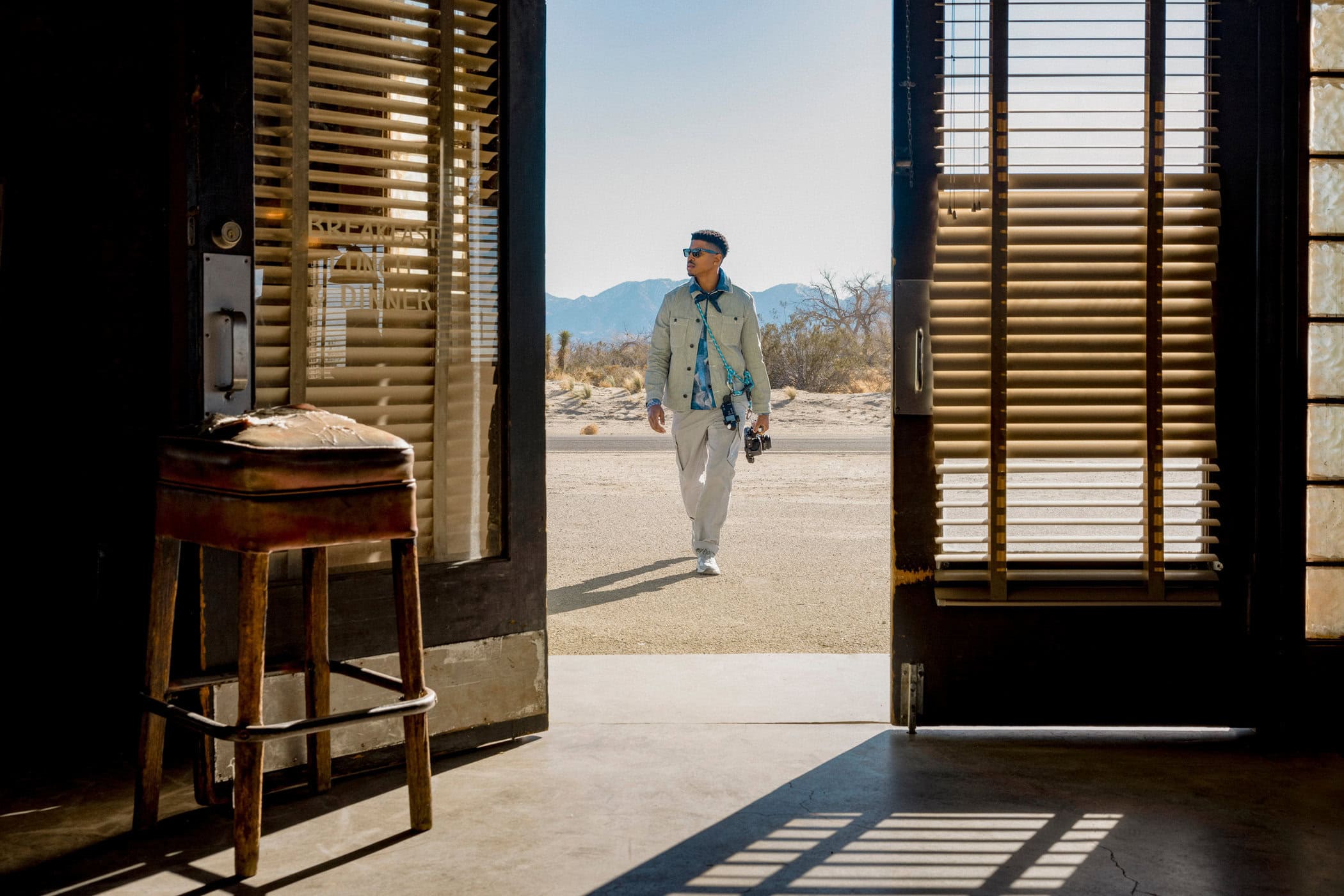 Adult male in a beige jacket walks out of a building with blinds into a desert landscape on a sunny day with mountains in the distance.