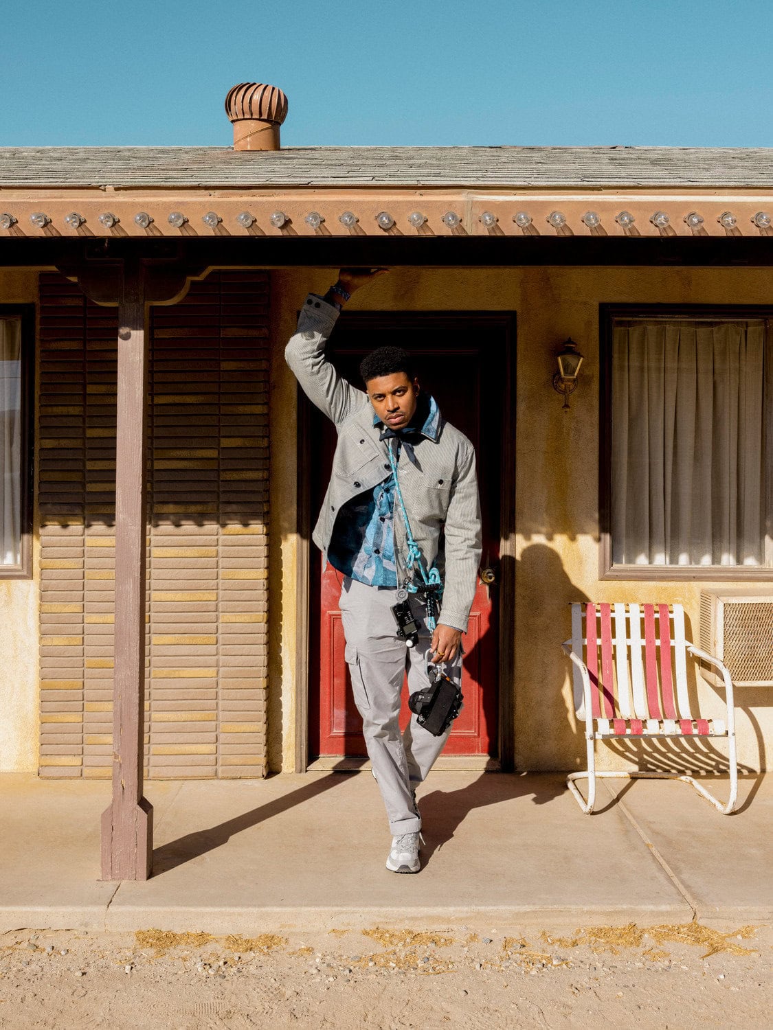A stylish adult man poses in front of a motel wearing a gray jacket, blue shirt, and light gray pants on a sunny day outdoors.