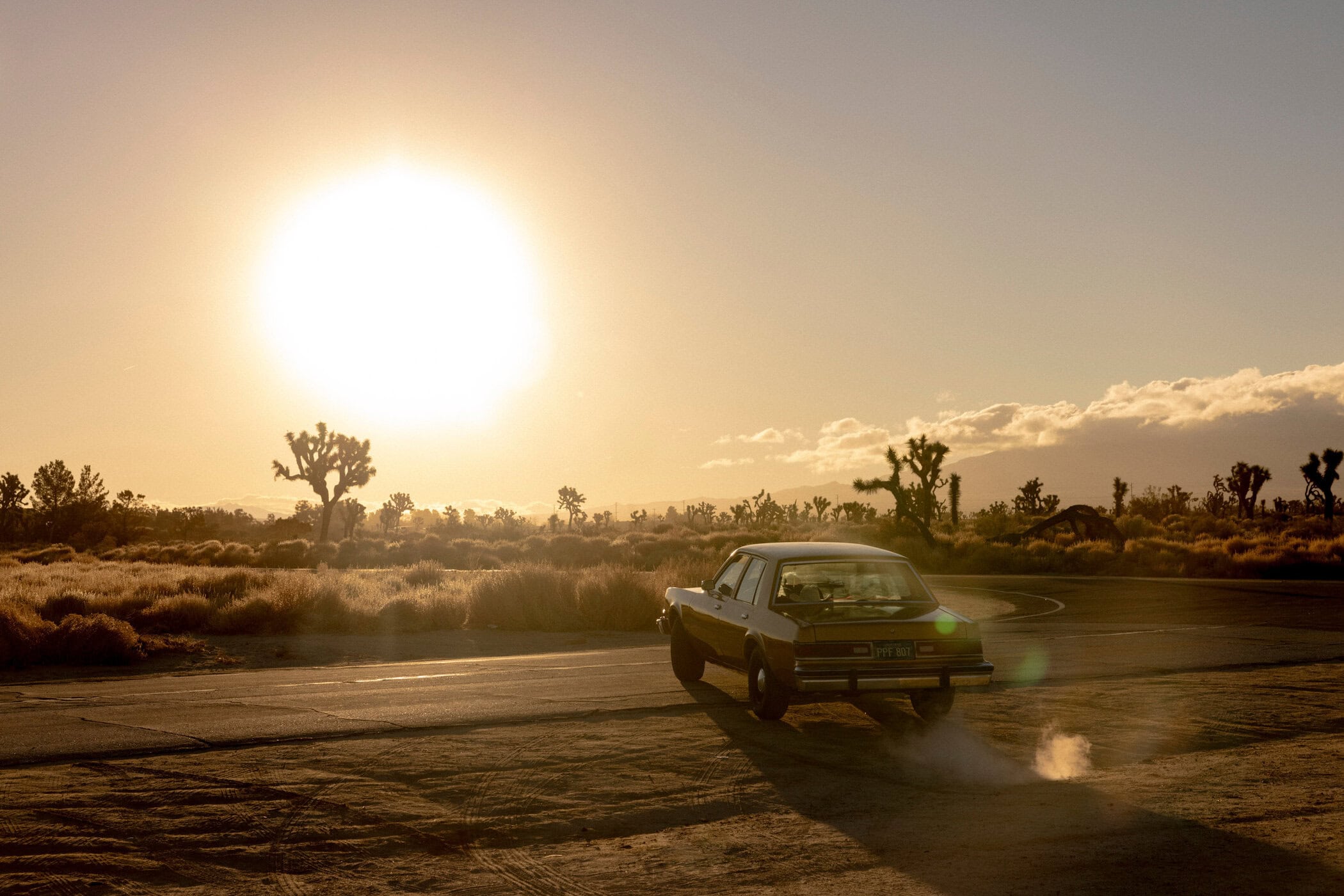 Vintage car driving into a desert sunset with Joshua trees in the background, creating a scenic and nostalgic image.