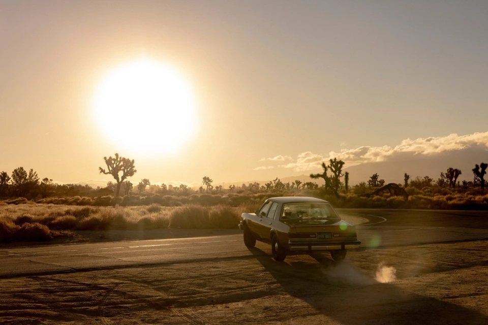 Vintage car driving into a desert sunset with Joshua trees in the background, creating a scenic and nostalgic image.