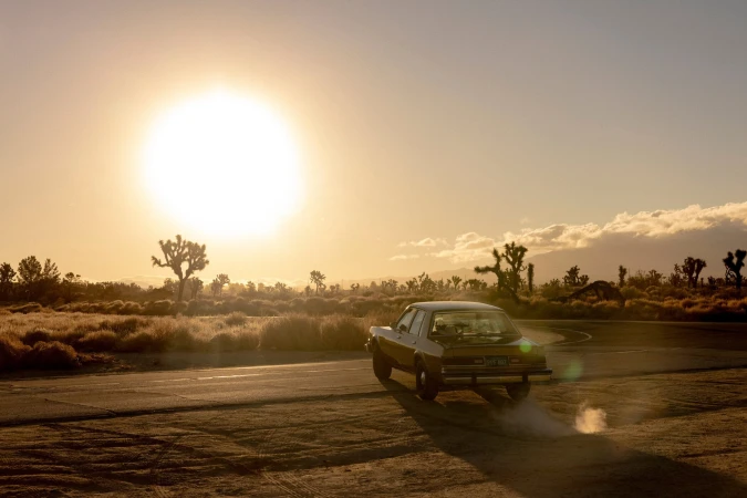 Vintage car driving into a desert sunset with Joshua trees in the background, creating a scenic and nostalgic image.