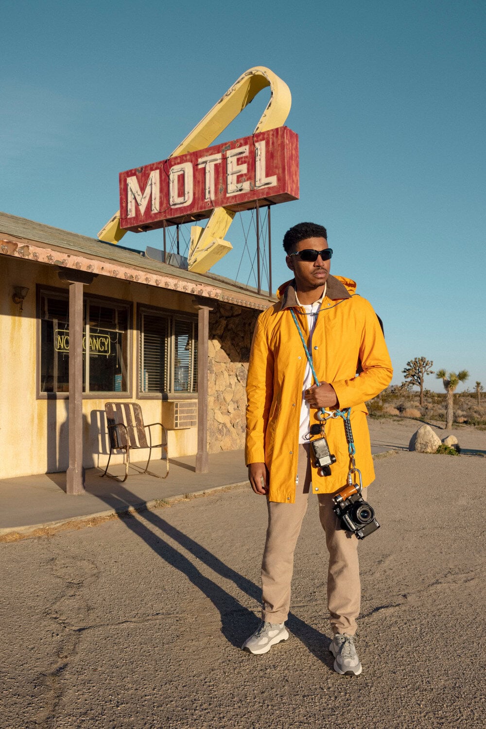 Adult male in yellow jacket standing in front of motel sign, holding camera. Clear blue sky and desert landscape.