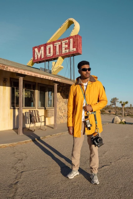 Adult male in yellow jacket standing in front of motel sign, holding camera. Clear blue sky and desert landscape.