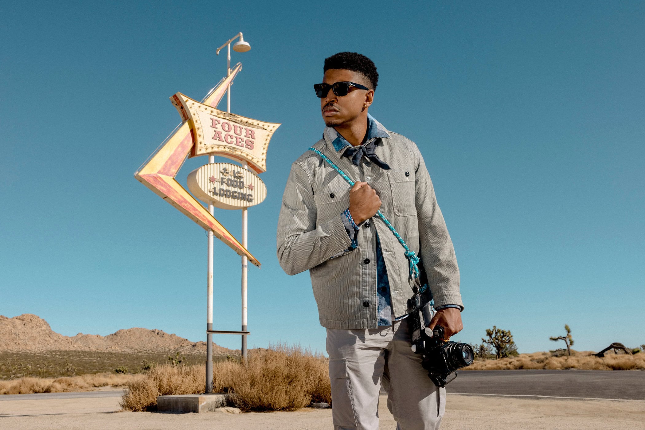 An adult male with a camera standing near a vintage motel sign on a desert landscape with blue skies. He's wearing casual attire.