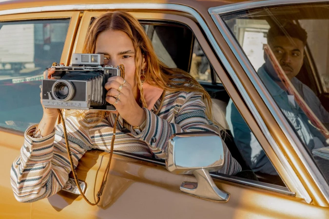 Woman photographs out the window of a vintage car in the daytime, with another person visible in the backseat.