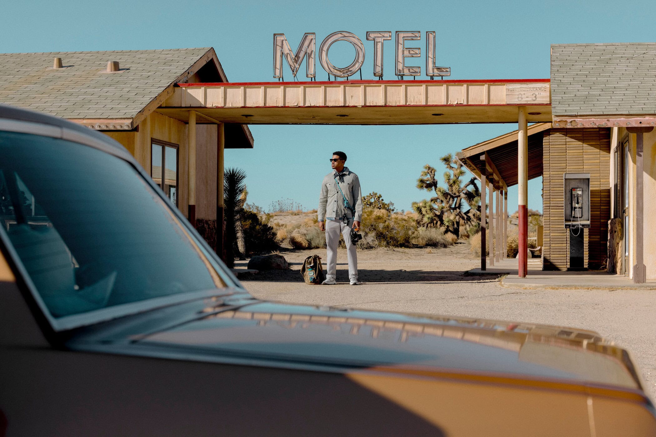 A man standing in front of an old motel in a desert landscape, with a vintage car partially visible, under a bright sky.