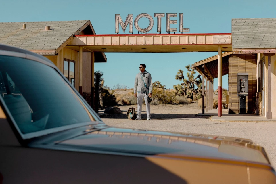 A man standing in front of an old motel in a desert landscape, with a vintage car partially visible, under a bright sky.