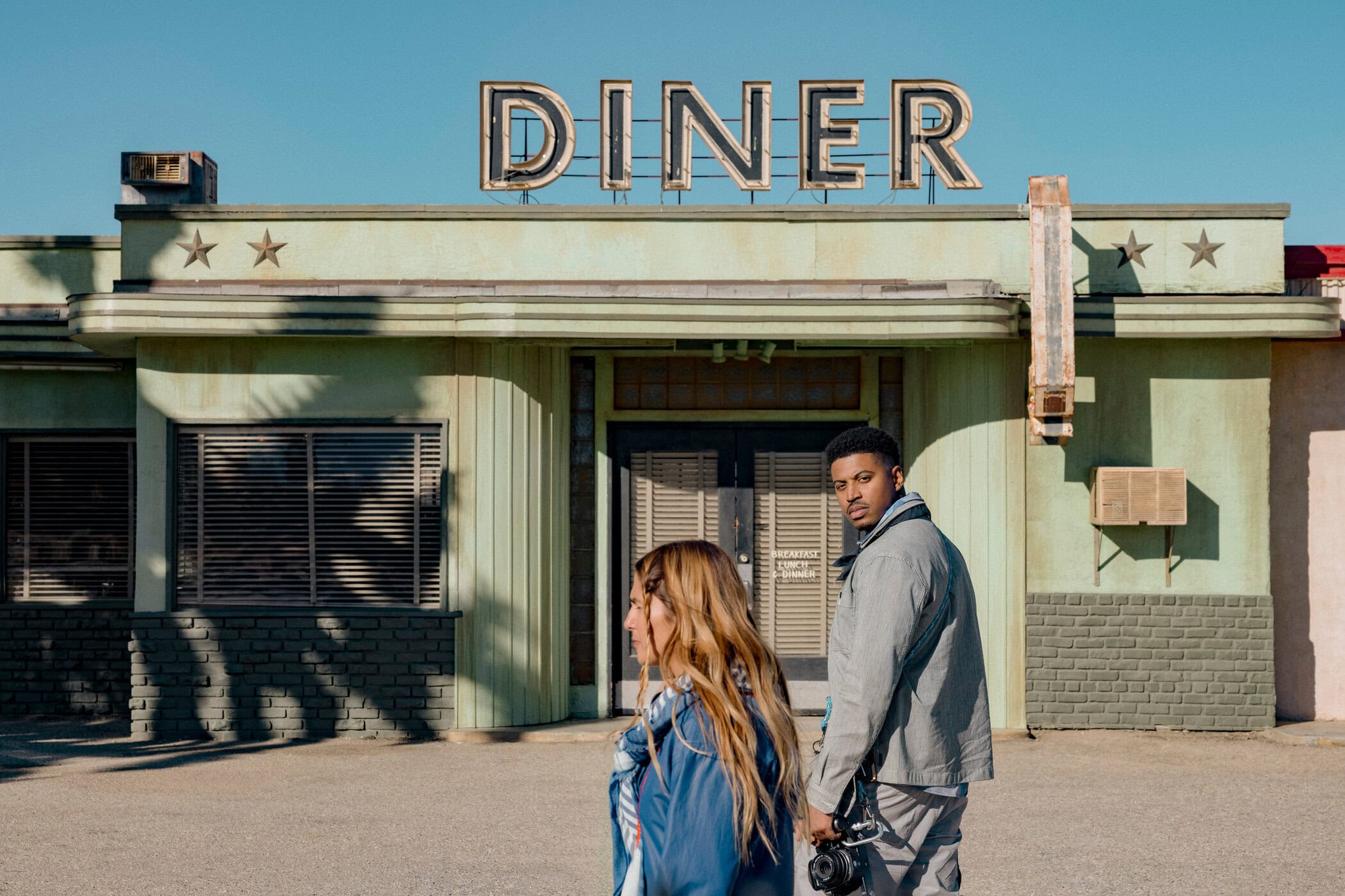 Two people stand in front of an old diner with a vintage sign on a sunny day, the man holding a camera, in a casual outdoor setting.