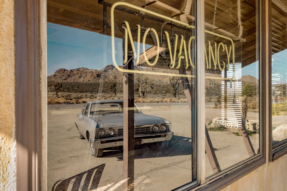 Vintage car parked near a motel in a desert landscape, seen through a window with a 'NO VACANCY' neon sign, under a clear blue sky.