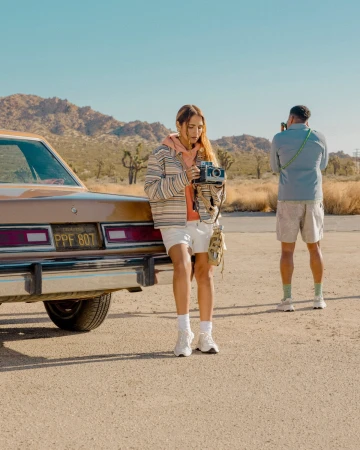 Woman holding vintage camera leaning against a classic car in a desert landscape with man photographing