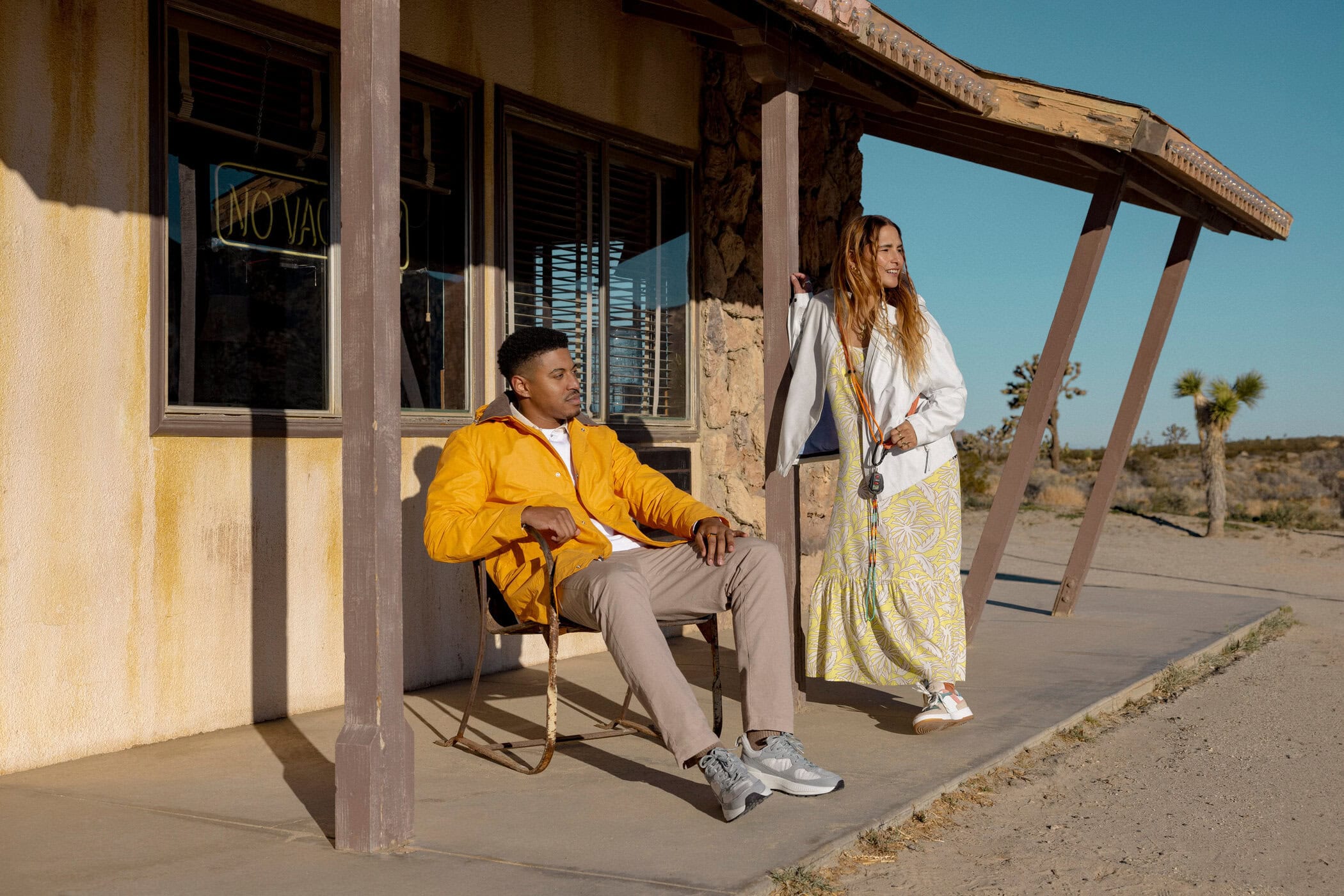 A man sits in a chair outside an old building while a woman stands nearby in a desert landscape under a clear blue sky.