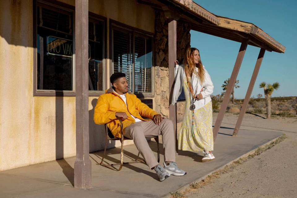 A man sits in a chair outside an old building while a woman stands nearby in a desert landscape under a clear blue sky.