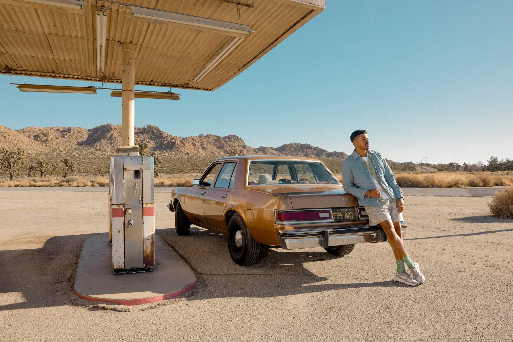 Man leans on vintage car at desert gas station under canopy, wearing light-colored shorts and shirt, mountains in background, sunny day.