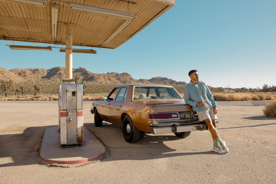 Man leans on vintage car at desert gas station under canopy, wearing light-colored shorts and shirt, mountains in background, sunny day.