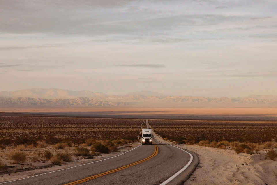 Semi-truck drives along asphalt road through desert landscape with mountains and cloudy sky, under soft, neutral-toned lighting.