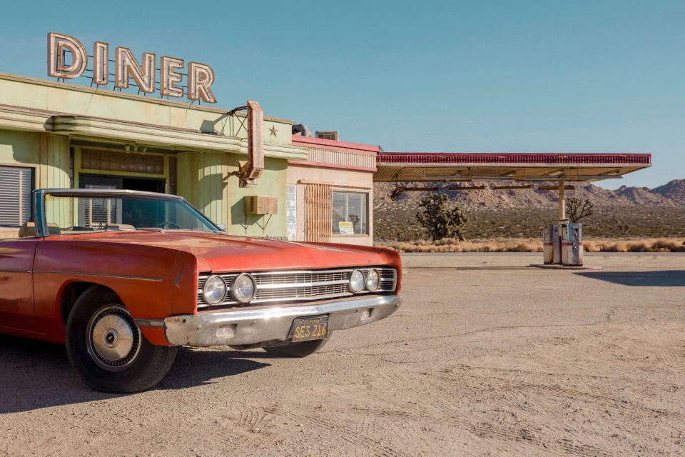 Vintage red convertible car parked outside a retro diner and gas station in a desert landscape under a clear blue sky, classic American scene.