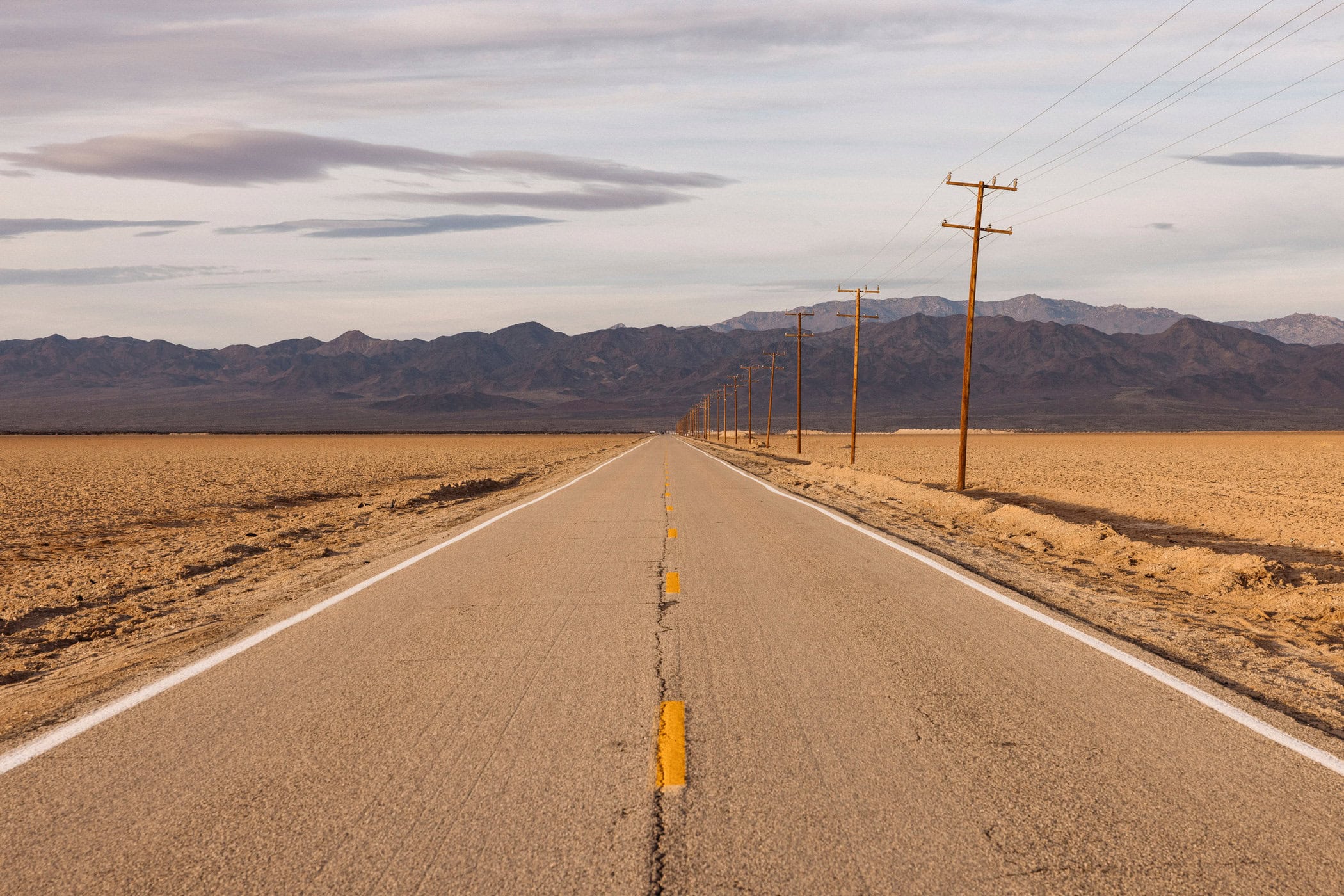 Straight desert highway under a cloudy sky with distant mountains and wooden utility poles under soft light, conveying a sense of solitude.