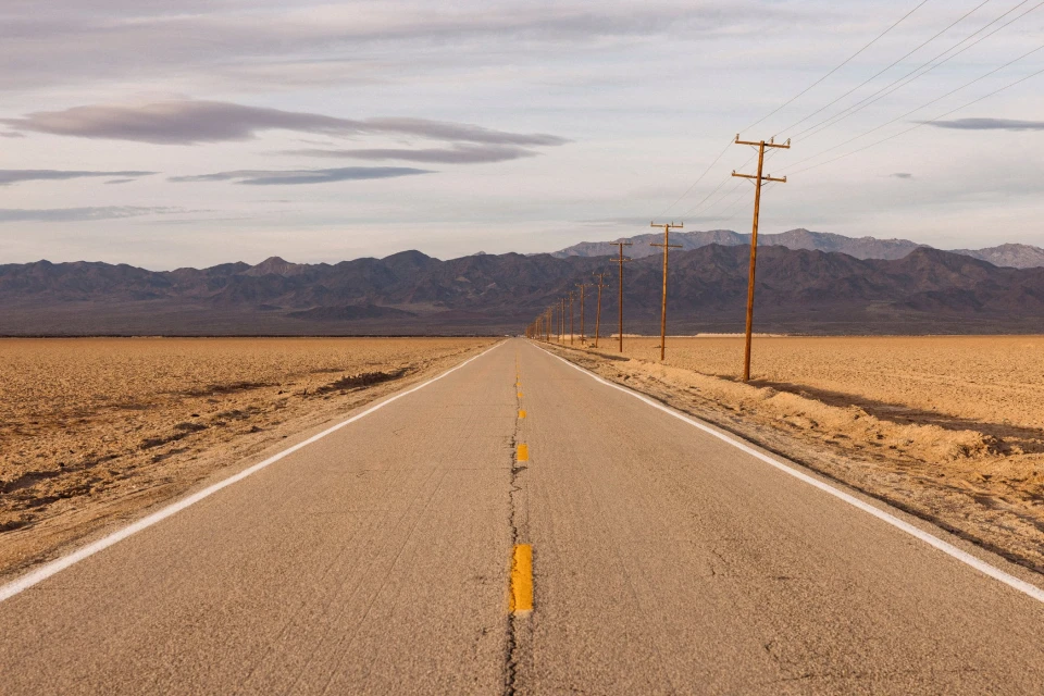 Straight desert highway under a cloudy sky with distant mountains and wooden utility poles under soft light, conveying a sense of solitude.