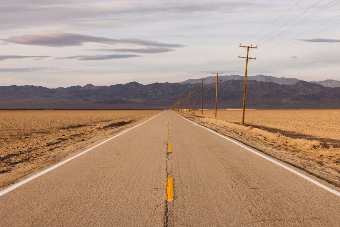 Straight desert highway under a cloudy sky with distant mountains and wooden utility poles under soft light, conveying a sense of solitude.