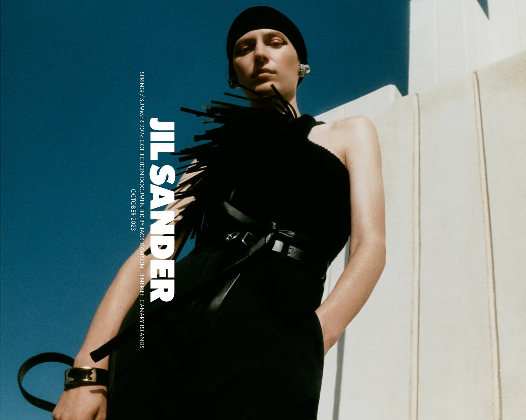 Fashion editorial shot of a woman in a black dress and cap posing against a bright blue sky and a white building on a sunny day.