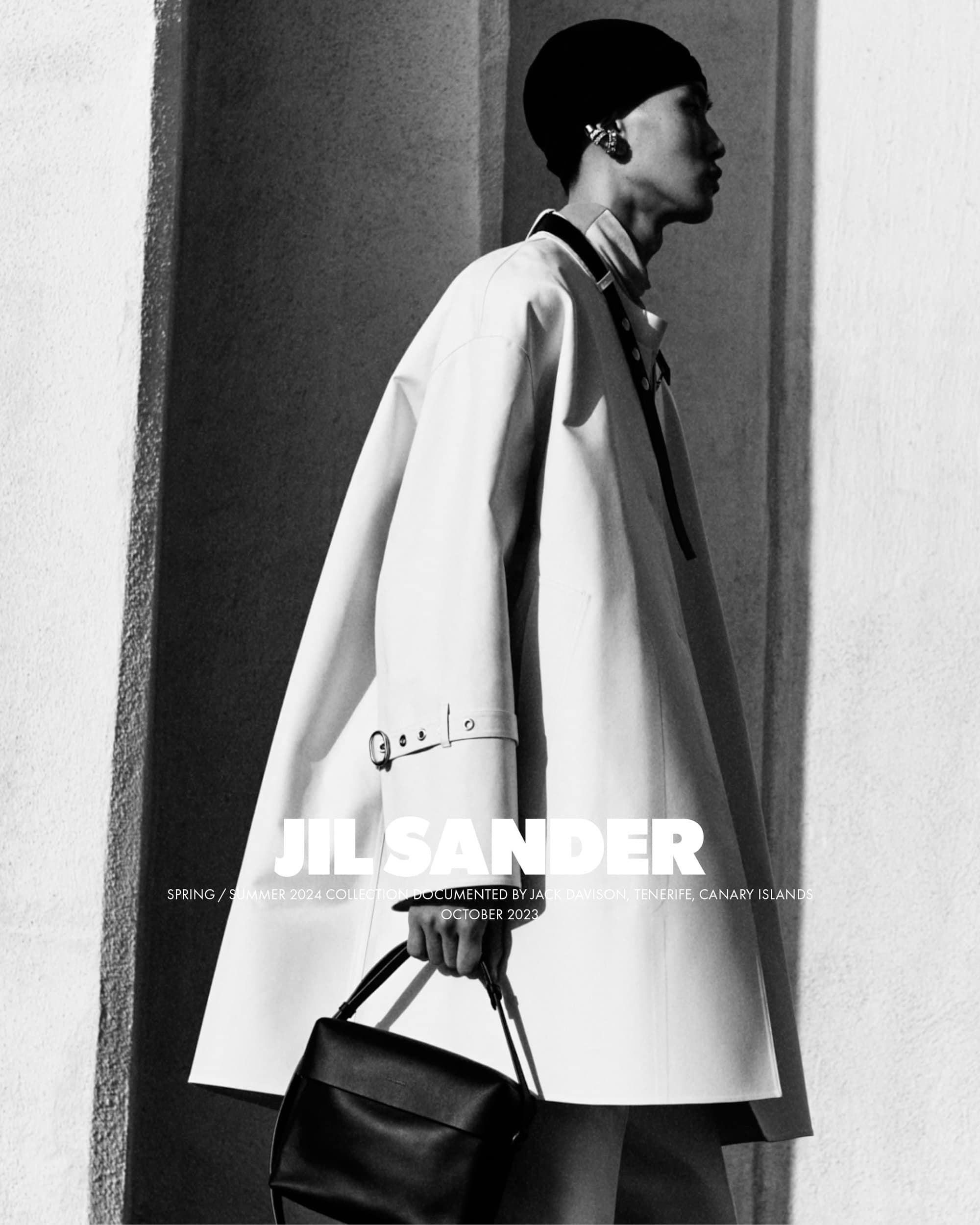 Black and white fashion shot of a person wearing a white coat and holding a black handbag near a textured wall in bright daylight.