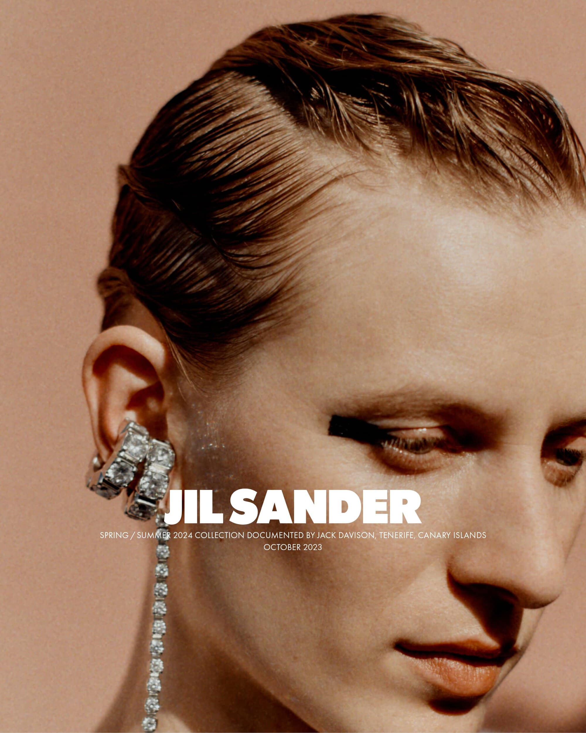 Close-up fashion shot of a female model with wet-look hair and a large crystal earring, wearing black eyeliner and posing against a warm background.
