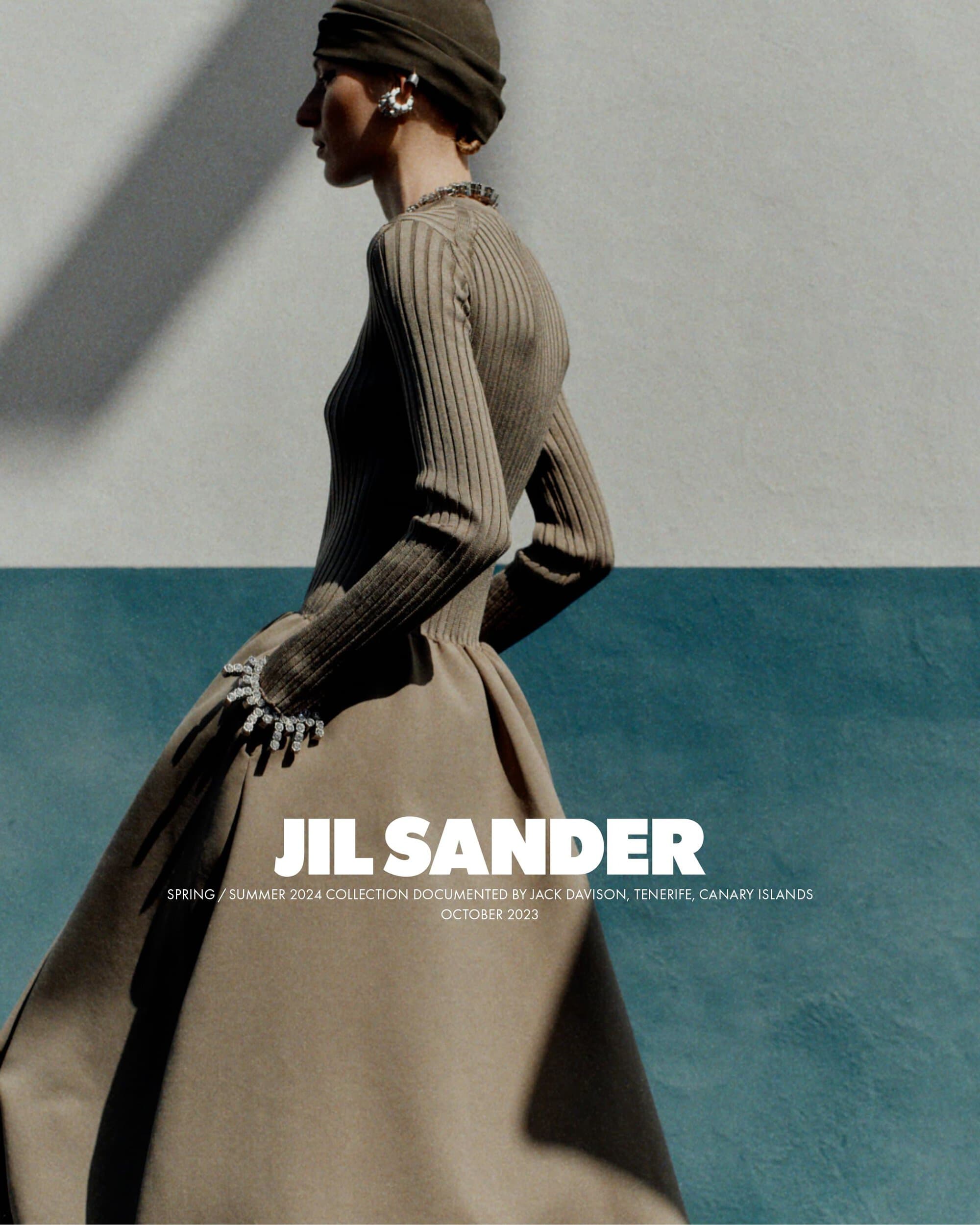 Fashion editorial shot of a woman in a long-sleeved top, olive turban, large earrings, and tan dress against a blue-and-white wall.