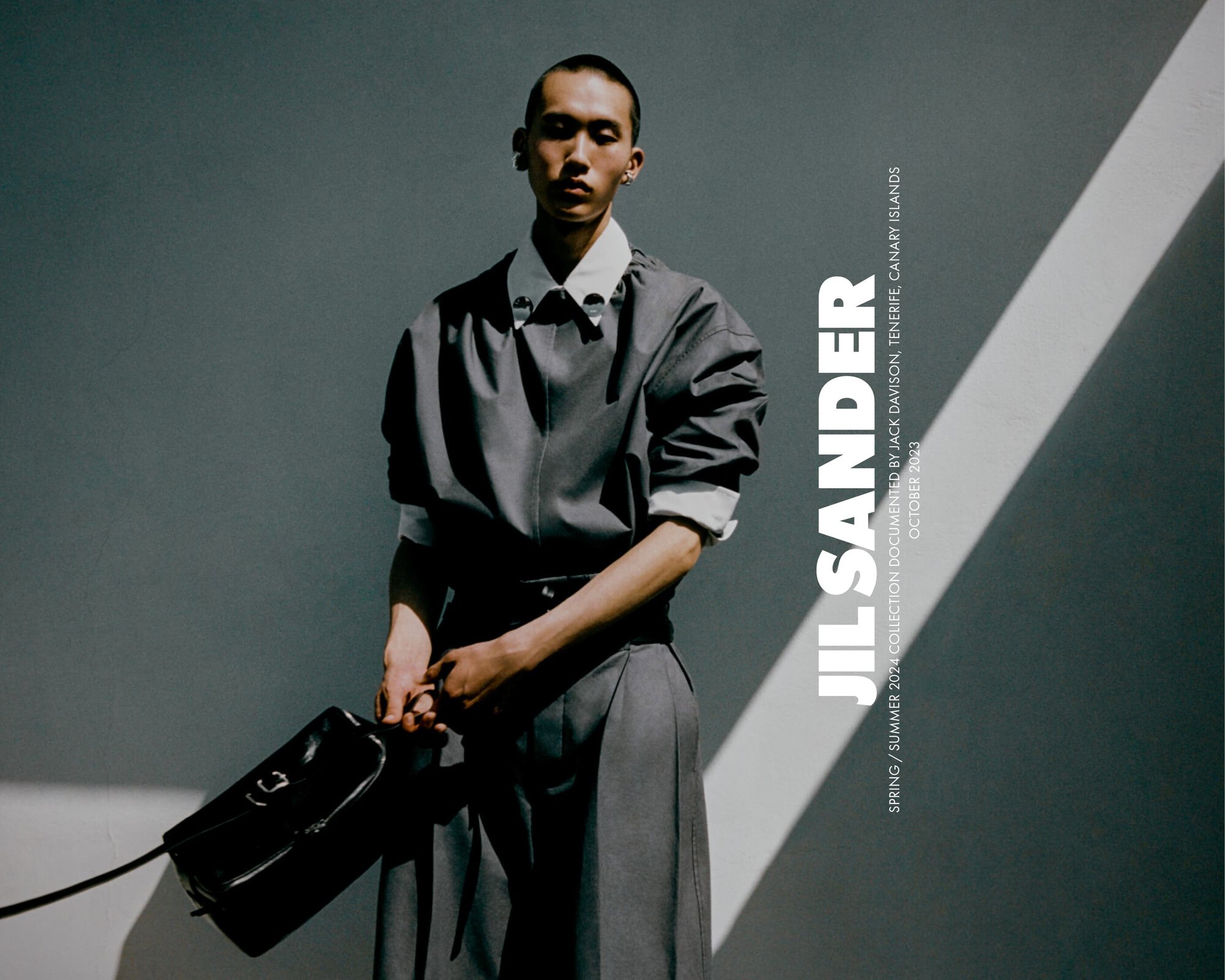 An adult male with short hair and grey clothing holds a black bag in front of a grey wall with a bright light, studio shot.