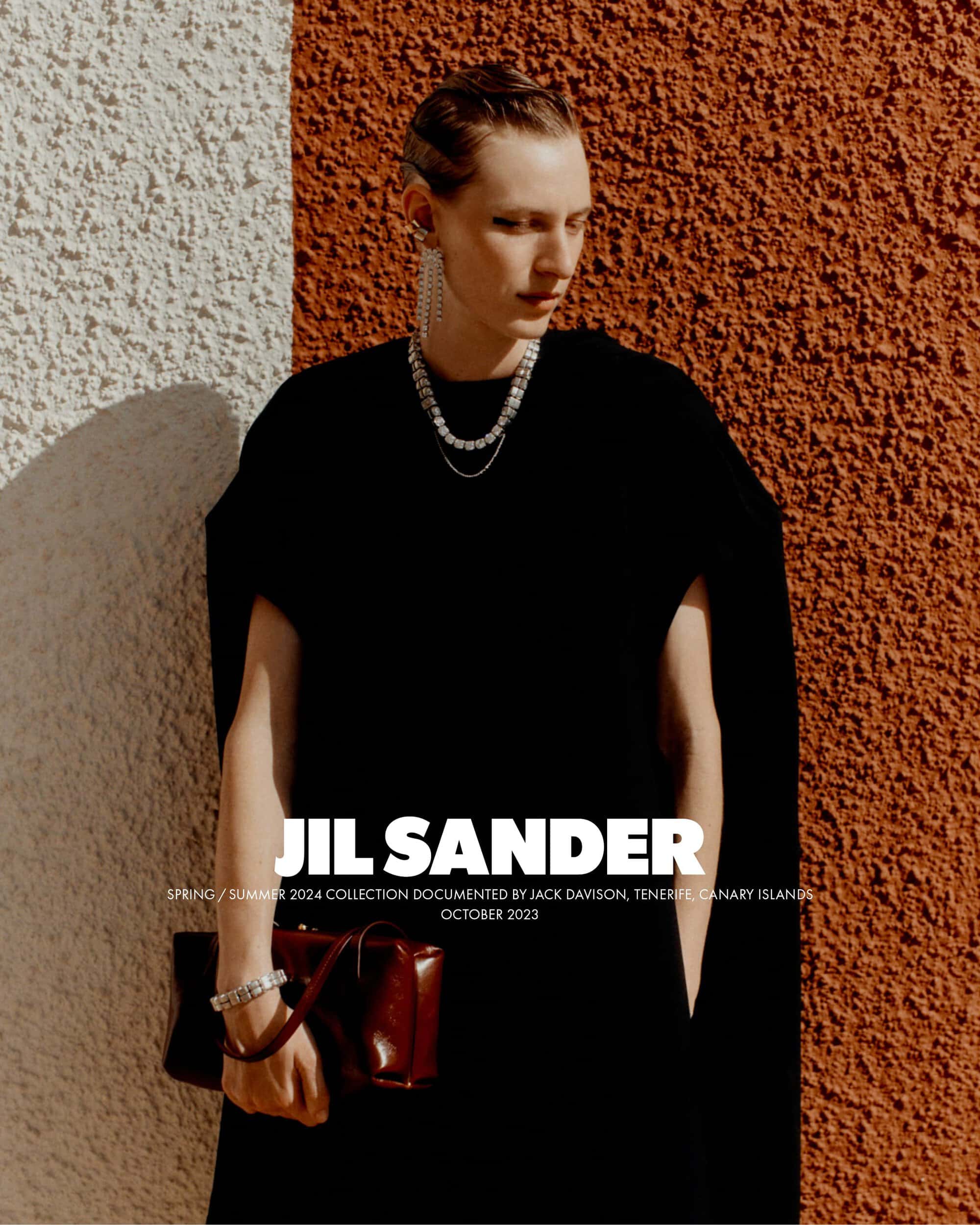 Fashion editorial shot of an adult female model in a black dress holding a purse, posing against a two-tone textured wall.