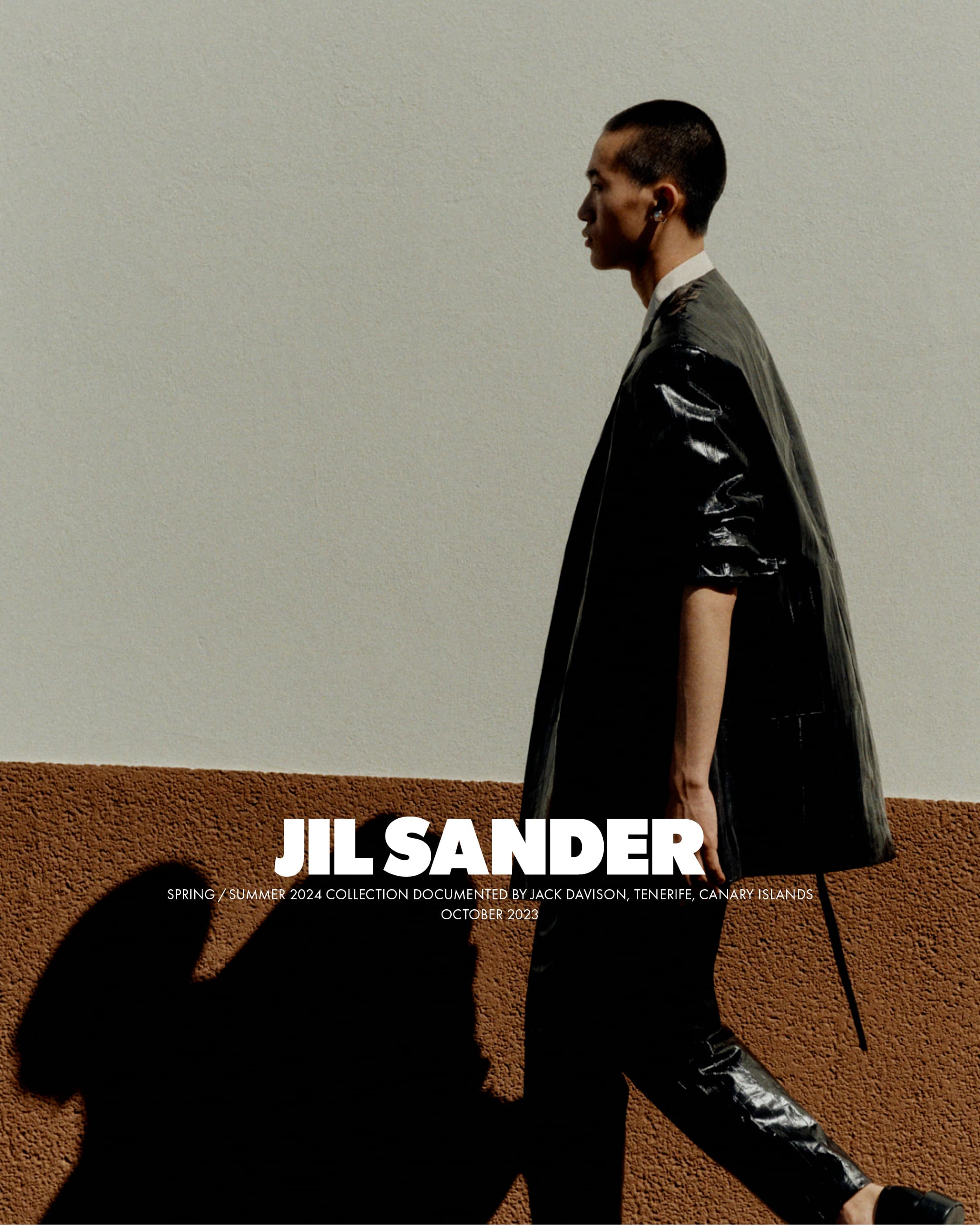 A stylish Asian male model showcases a black Jil Sander coat and pants while walking against a textured cream and brown wall in natural light.