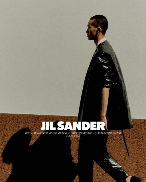A stylish Asian male model showcases a black Jil Sander coat and pants while walking against a textured cream and brown wall in natural light.