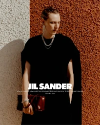 Model in black dress wearing crystal jewelry and holding a red handbag against a textured wall for Jil Sander Spring 2024 campaign