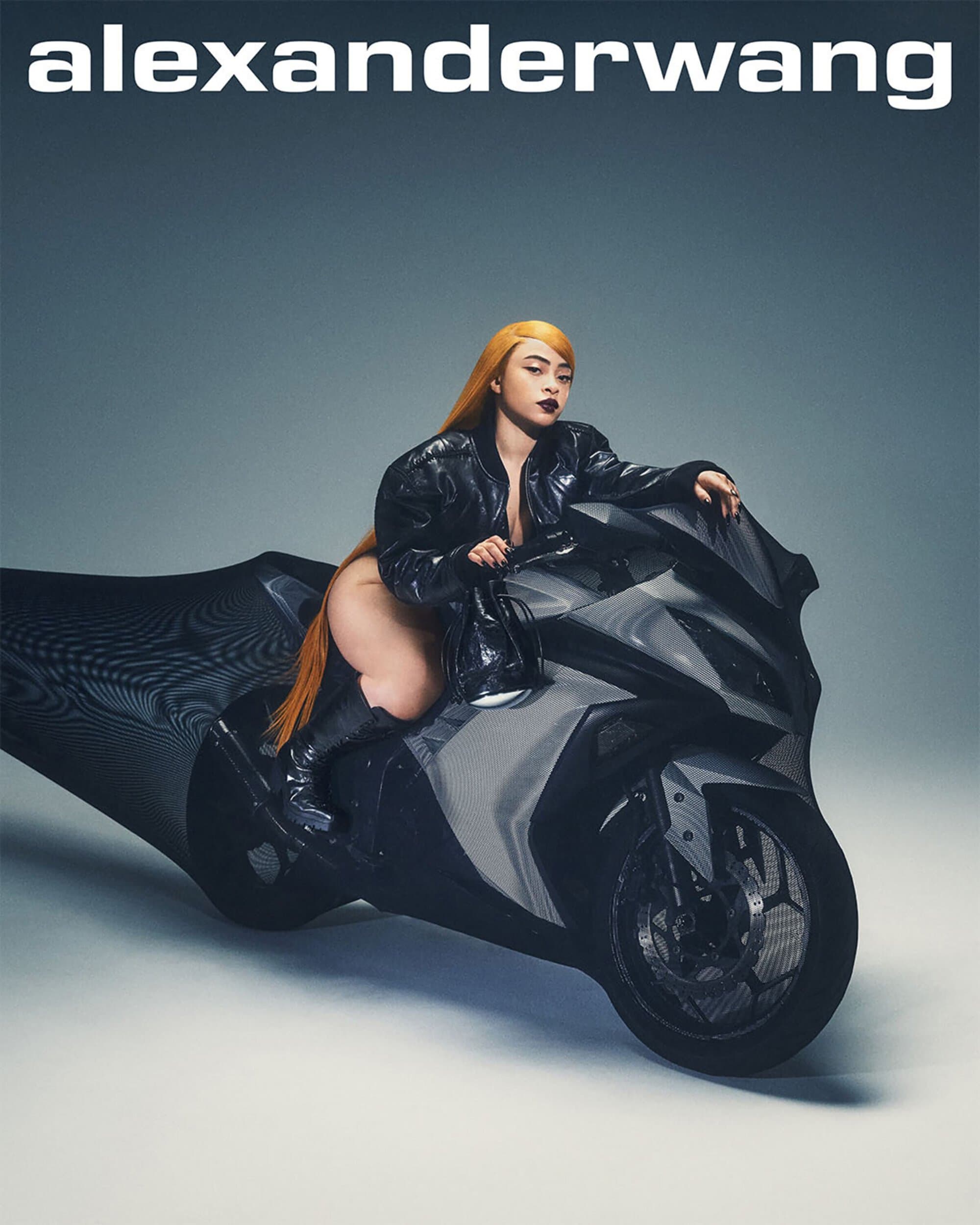 Adult woman with long orange hair poses seated on a black and silver motorcycle in a studio environment for a fashion editorial.