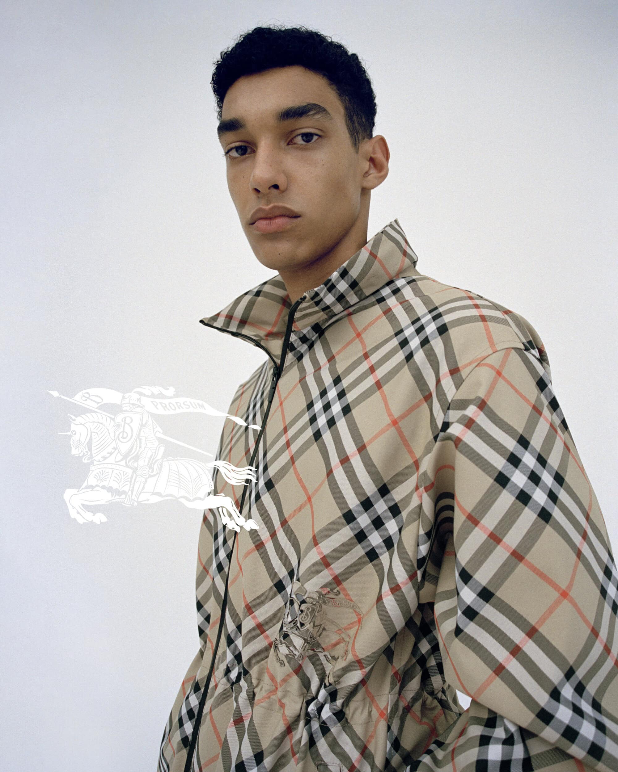 Fashion shot of a young adult male wearing a tan plaid jacket with a white logo on a white backdrop in a studio setting.