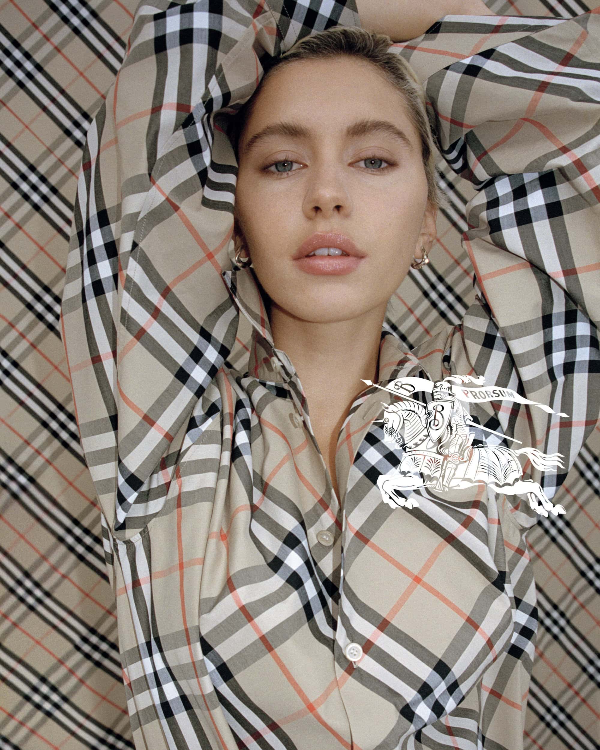Close-up studio shot of a woman in a tan plaid button-down, posing with a Burberry logo graphic overlay against a matching backdrop.
