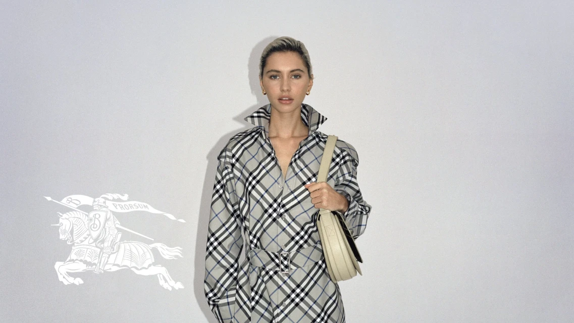 Blonde woman posing in a grey and black plaid Burberry shirt dress with a beige bag, standing against a neutral studio backdrop.