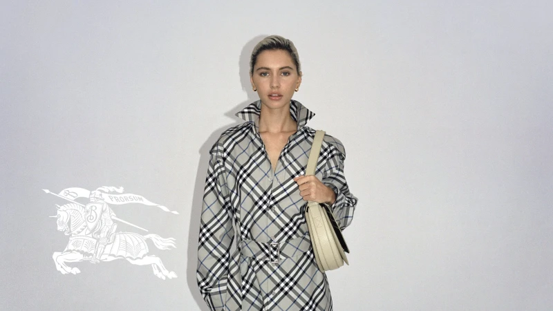 Blonde woman posing in a grey and black plaid Burberry shirt dress with a beige bag, standing against a neutral studio backdrop.