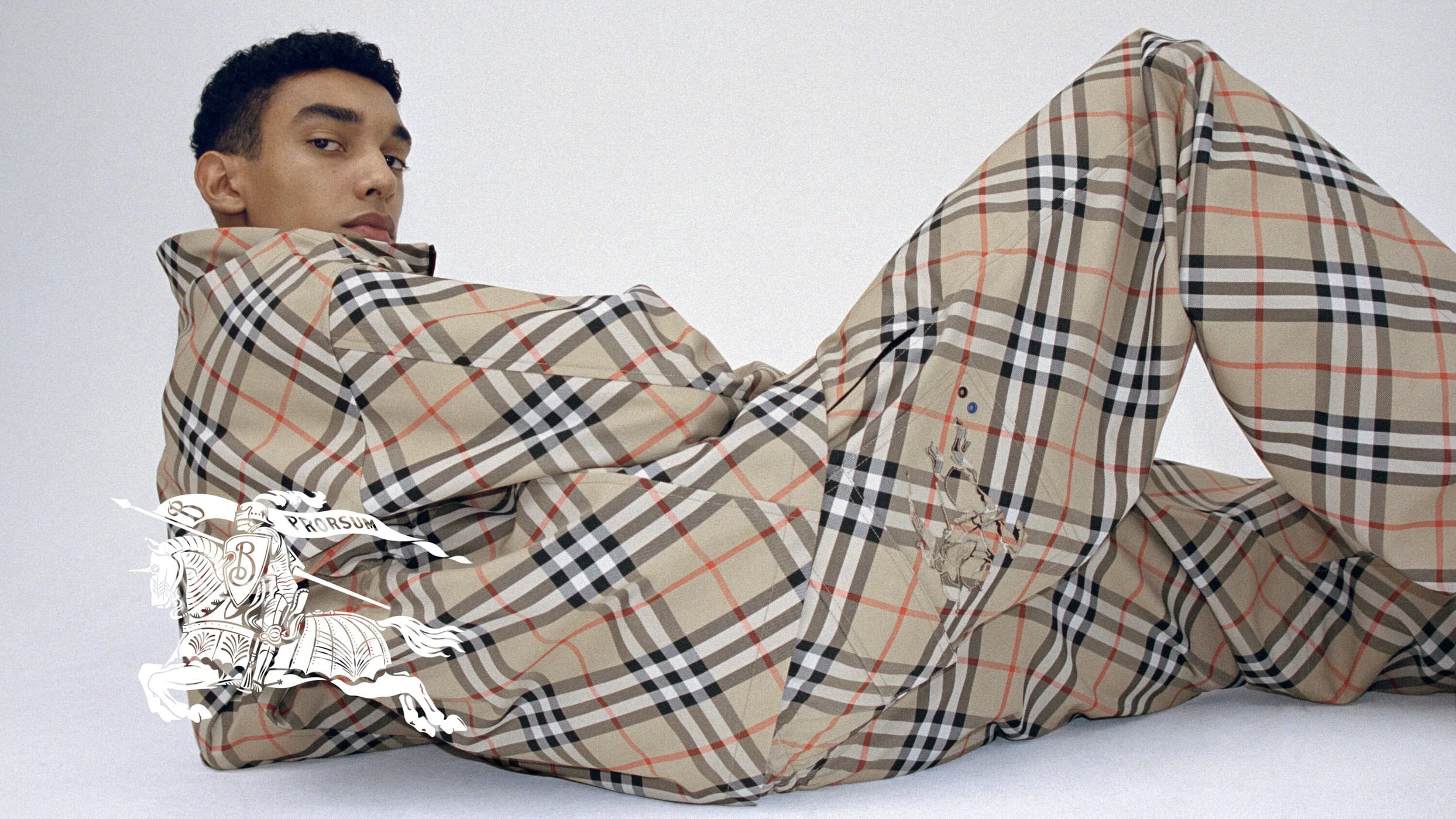 An adult male model lies on a white surface wearing a beige and black plaid patterned shirt and pants; Burberry logo illustration.