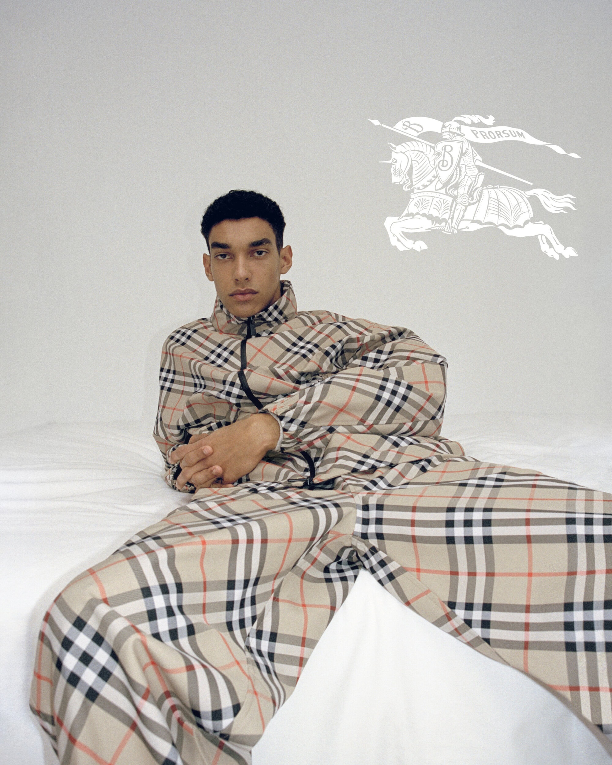 Adult male laying on a bed wearing a Burberry check tracksuit with Burberry logo visible in the background on a plain white wall.