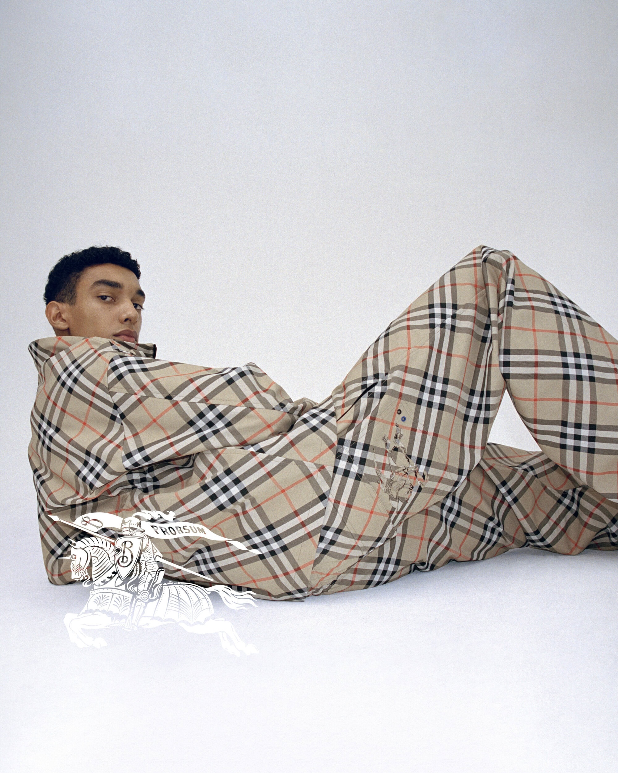Adult male model in a studio wearing a Burberry print pajama set, lying on his side, and looking at the camera in a studio setting.