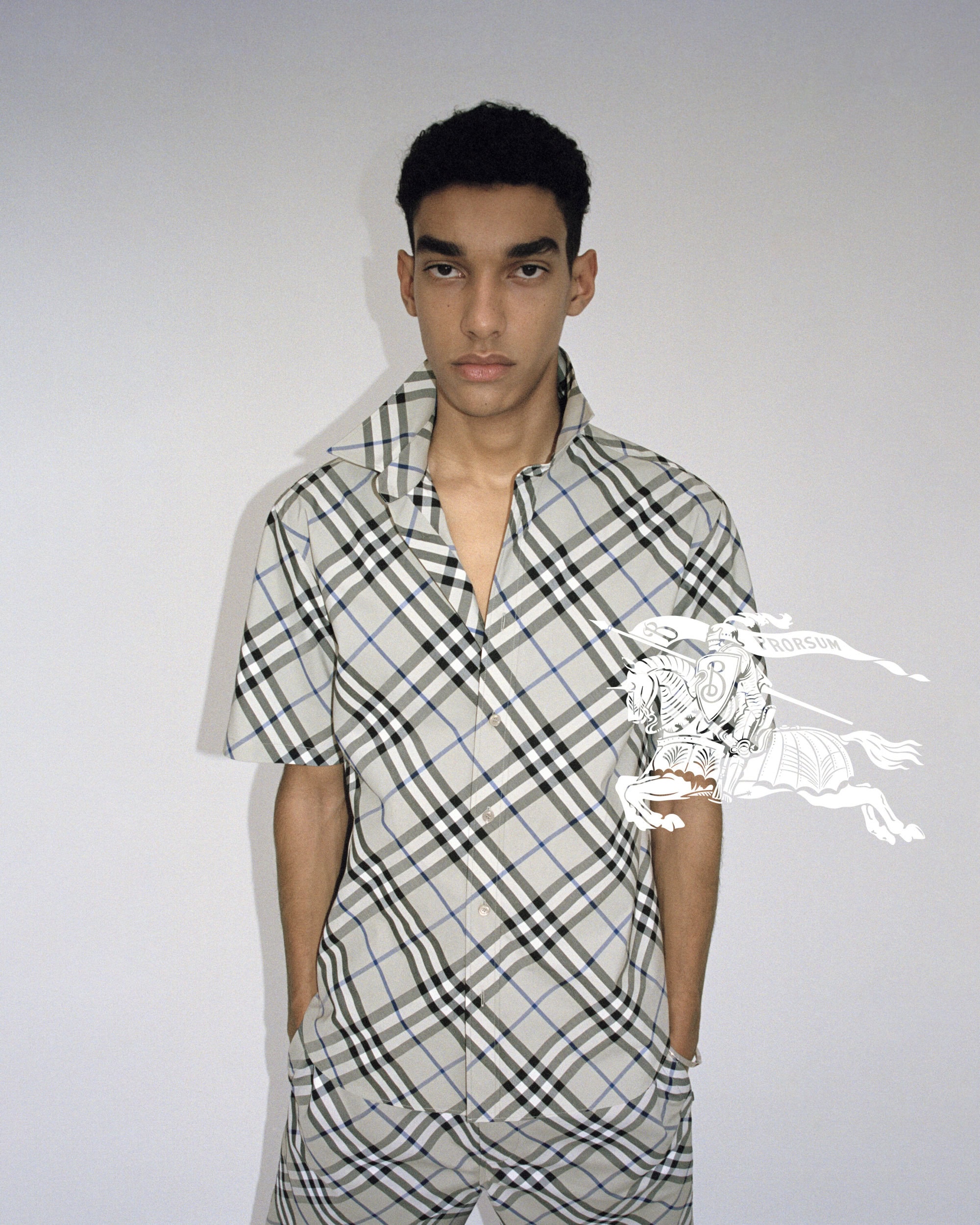 Young adult male poses in a grey plaid shirt and pants with short dark hair. The background is a neutral off-white in a portrait shot.