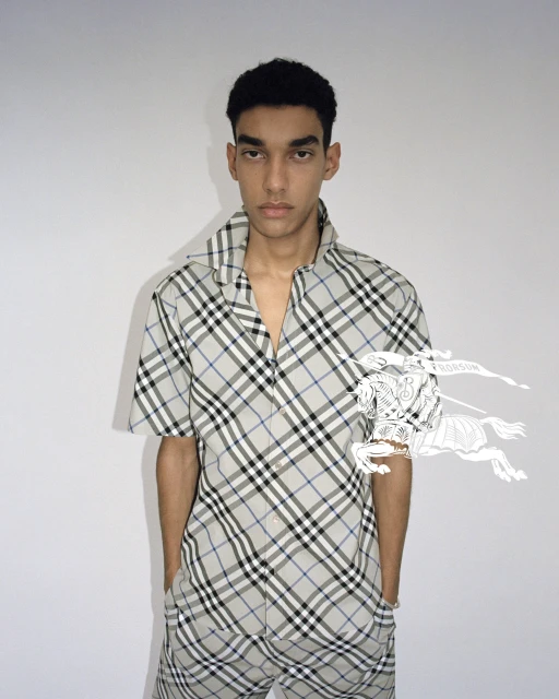 Young adult male poses in a grey plaid shirt and pants with short dark hair. The background is a neutral off-white in a portrait shot.