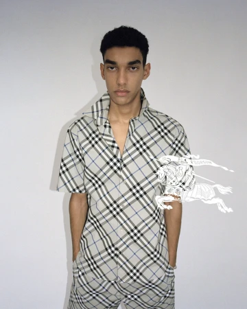 Young adult male poses in a grey plaid shirt and pants with short dark hair. The background is a neutral off-white in a portrait shot.